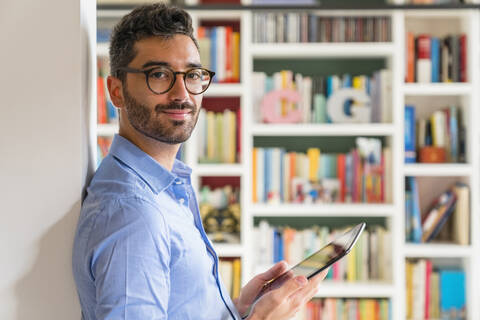 Portrait of smiling young man standing in front of bookshelves at home with digital tablet stock photo