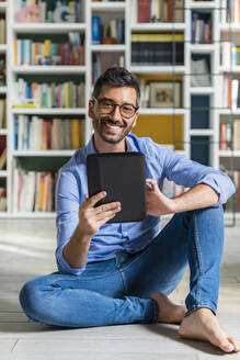 Porträt eines barfüßigen jungen Mannes, der vor einem Bücherregal auf dem Boden sitzt und ein digitales Tablet benutzt - MGIF00851