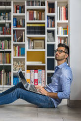 Porträt eines nachdenklichen jungen Mannes, der vor einem Bücherregal auf dem Boden sitzt und einen Laptop benutzt - MGIF00852