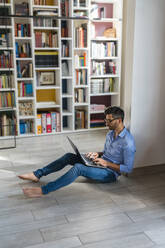 Barfüßiger junger Mann sitzt zu Hause auf dem Boden und benutzt einen Laptop - MGIF00854