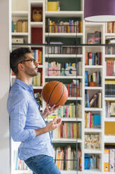 Nachdenklicher junger Mann mit Basketball vor einem Bücherregal zu Hause stehend - MGIF00859