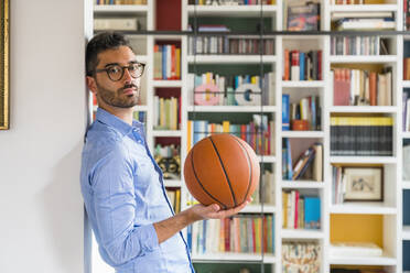 Portrait of young man with basketball standing in front of bookshelves at home - MGIF00860