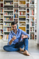 Porträt eines lächelnden jungen Mannes, der zu Hause auf dem Boden vor einem Bücherregal sitzt und auf sein Smartphone schaut - MGIF00861