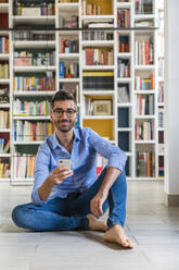 Porträt eines lächelnden jungen Mannes, der mit seinem Smartphone vor einem Bücherregal auf dem Boden sitzt - MGIF00862