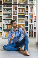 Porträt eines nachdenklichen jungen Mannes mit Smartphone, der zu Hause vor einem Bücherregal auf dem Boden sitzt und in die Ferne schaut - MGIF00863