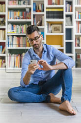 Porträt eines lächelnden jungen Mannes, der zu Hause vor einem Bücherregal auf dem Boden sitzt und ein Smartphone benutzt - MGIF00864