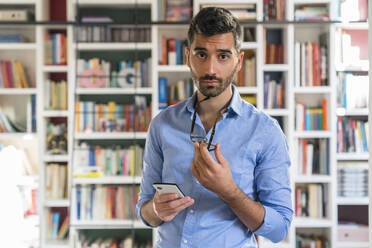 Portrait of sceptical young man with smartphone standing in front of bookshelves - MGIF00867