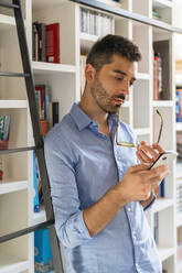Porträt eines nachdenklichen jungen Mannes, der vor einem Bücherregal steht und auf sein Mobiltelefon schaut - MGIF00869