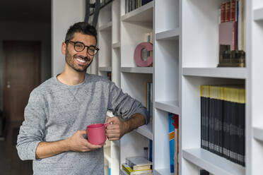 Porträt eines lächelnden jungen Mannes mit Tasse, der zu Hause vor einem Bücherregal steht - MGIF00873