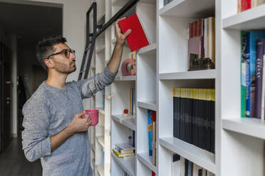 Porträt eines jungen Mannes mit Tasse, der zu Hause vor einem Bücherregal steht und ein Buch zur Hand nimmt - MGIF00874
