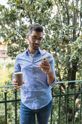 Portrait of young man standing on balcony with coffee to go looking at cell phone - MGIF00875