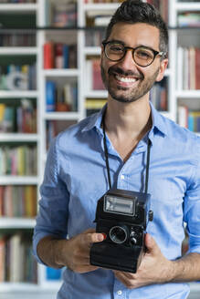 Porträt eines glücklichen jungen Mannes mit Sofortbildkamera vor einem Bücherregal - MGIF00878
