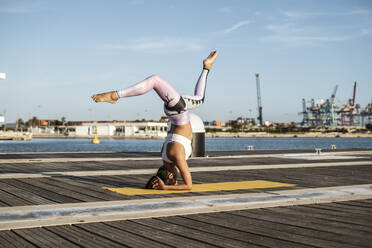 Asiatische Frau übt Yoga auf einem Pier am Hafen, Kopfstand mit Spagat - RCPF00093