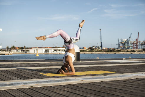 Asiatische Frau übt Yoga auf einem Pier am Hafen, Kopfstand mit Spagat - RCPF00093