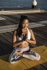 Asian woman practicing yoga on a pier at harbour - RCPF00096