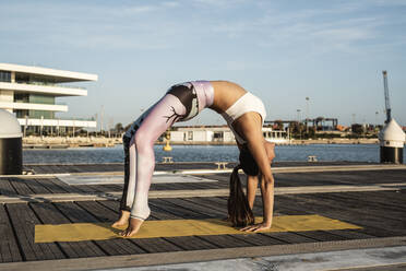 Asiatische Frau übt Yoga auf einem Pier am Hafen, nach oben gerichtet - RCPF00099