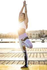 Asiatische Frau übt Yoga auf einem Pier am Hafen, Baumposition - RCPF00103