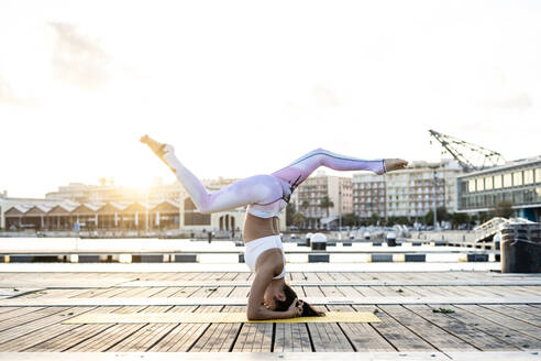 Asiatische Frau übt Yoga auf einem Pier am Hafen bei Sonnenuntergang, Kopfstand mit Spagat - RCPF00116