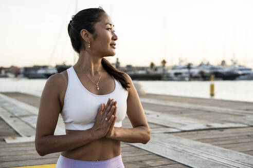 Asian woman practicing yoga on a pier at harbour in the evening light, prayer position - RCPF00122