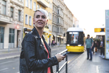 Porträt einer blonden Frau mit Smartphone an einer Straßenbahnhaltestelle, Berlin, Deutschland - WPEF02248