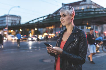 Porträt einer blonden Frau, die abends am Straßenrand steht und ein Mobiltelefon benutzt, Berlin, Deutschland - WPEF02252