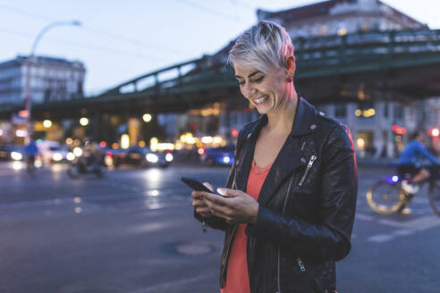 Porträt einer lachenden blonden Frau, die abends am Straßenrand steht und ein Mobiltelefon benutzt, Berlin, Deutschland - WPEF02253