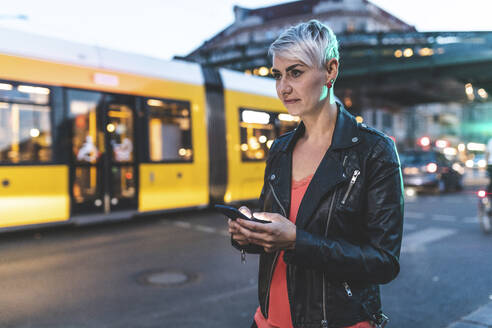 Porträt einer blonden Frau, die abends am Straßenrand steht und ein Mobiltelefon benutzt, Berlin, Deutschland - WPEF02254