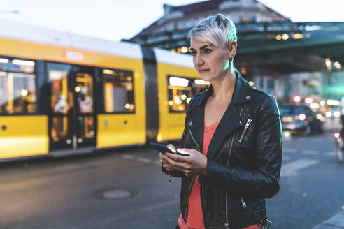 Portrait of blond woman standing at roadside in the evening using mobile phone, Berlin, Germany - WPEF02254