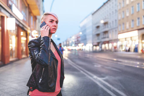 Porträt einer blonden Frau, die am Abend am Straßenrand steht und telefoniert, Berlin, Deutschland - WPEF02255
