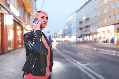 Portrait of blond woman on the phone standing at roadside in the evening, Berlin, Germany - WPEF02255