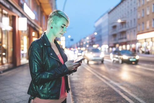 Blond woman standing at roadside in the evening using mobile phone, Berlin, Germany - WPEF02256