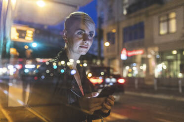 Porträt einer blonden Frau an einer abendlichen Straßenbahnhaltestelle, die ein Mobiltelefon benutzt, Berlin, Deutschland - WPEF02258