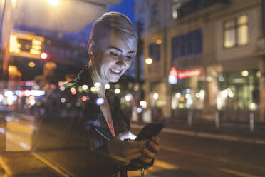 Porträt einer lächelnden blonden Frau an einer Straßenbahnhaltestelle, die auf ihr Mobiltelefon schaut, Berlin, Deutschland - WPEF02259