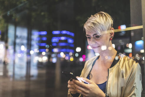 Porträt einer lächelnden blonden Frau, die in der nächtlichen Stadt ein Mobiltelefon benutzt, Berlin, Deutschland - WPEF02262