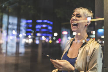 Porträt einer lachenden blonden Frau, die nachts ein Mobiltelefon benutzt, Berlin, Deutschland - WPEF02263