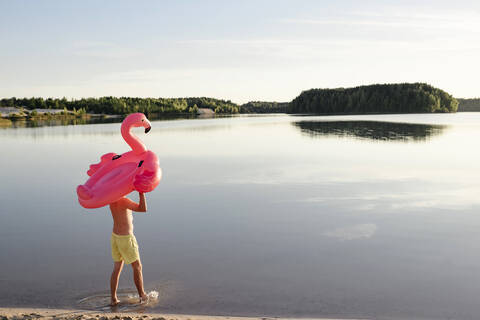 Junger Mann mit Flamingo-Poolschwimmer, der am Seeufer spazieren geht, lizenzfreies Stockfoto