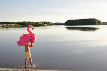 Young man with flamingo pool float walking at lakeshore - EYAF00647