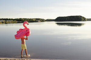 Junger Mann mit Flamingo-Poolschwimmer, der am Seeufer spazieren geht - EYAF00647