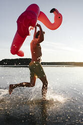 Young man running with flamingo pool float into a lake - EYAF00651