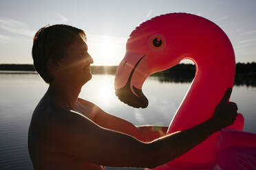 Junger Mann mit Flamingo-Poolschwimmer bei Sonnenuntergang - EYAF00652