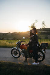 Junges Paar mit Oldtimer-Motorrad steht auf der Landstraße bei Sonnenuntergang und schaut auf die Aussicht, Toskana, Italien - JPIF00240