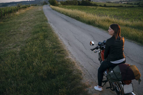 Junge Frau mit Oldtimer-Motorrad steht auf der Landstraße bei Sonnenuntergang mit Blick auf die Aussicht, Toskana, Italien - JPIF00244