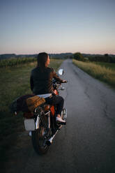 Rückenansicht einer jungen Frau, die bei Sonnenuntergang auf einem Motorrad sitzt und die Aussicht genießt, Toskana, Italien - JPIF00245