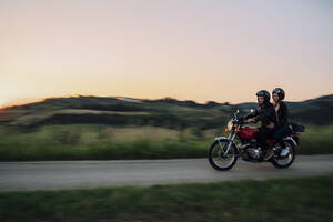 Junges Paar fährt mit einem alten Motorrad auf einer Landstraße bei Sonnenuntergang, Toskana, Italien - JPIF00250