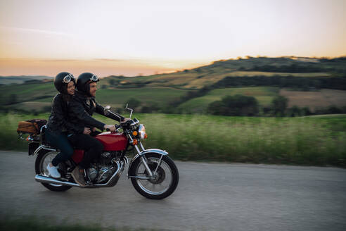 Junges Paar fährt mit einem alten Motorrad auf einer Landstraße bei Sonnenuntergang, Toskana, Italien - JPIF00251