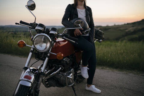 Crop shot of young woman with vintage motorbike on country road at sunset, Tuscany, Italy stock photo