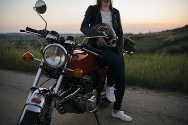 Crop shot of young woman with vintage motorbike on country road at sunset, Tuscany, Italy - JPIF00255