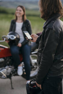 Junger Motorradfahrer steht auf der Landstraße und benutzt sein Smartphone, während seine Freundin im Hintergrund wartet - JPIF00256