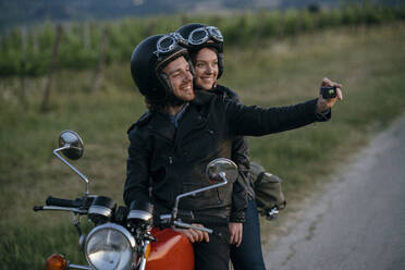 Portrait of happy young couple on vintage motorbike taking a selfie at roadside - JPIF00258