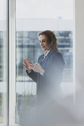 Reife Geschäftsfrau mit Handy am Fenster im Büro - MOEF02512